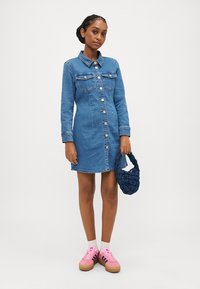 Denim obleka s ovratnikom, dolgimi rokavi, dvema žepoma na prsih in gumbom za zapiranje. Oblečena s rožnatimi supergami in strukturirano modro torbo.