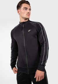 Zwarte zip-up sportjack met witte zijkanten, hoge kraag en geribbelde manchetten. Voorzien van een logo op de linkerborst en ritszakken.