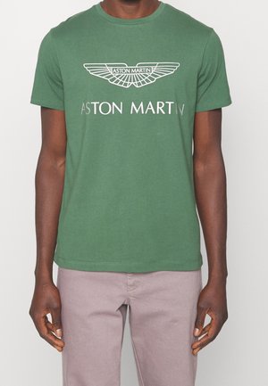 Person trägt grünes T-Shirt mit Aston Martin-Logo und helle Hose, steht vor schlichtem hellem Hintergrund.