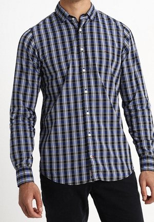 Hombre vistiendo una camisa de cuadros azul, negra y blanca de manga larga y con botones, con pantalones negros, de pie frente a un fondo blanco.