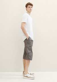Weißes T-Shirt mit kurzen Ärmeln, graue Cargo-Shorts mit Seitentaschen und hellfarbige Sneakers mit beigen Akzenten. Das Model steht im Profil.