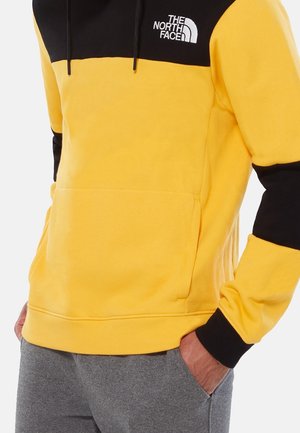 Person trägt einen gelb-schwarzen North Face Hoodie mit Fronttasche und graue Hose, Hände in Hosentaschen gesteckt.