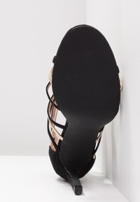Sandale noire à talon haut avec fines brides et semelle brillante, positionnée verticalement contre une surface d'angle blanche.