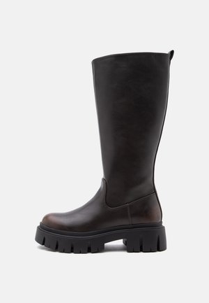 Botte haute noire en cuir avec une semelle épaisse et épaisse et un bout arrondi, vue de profil sur un fond clair uni.