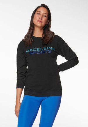 Schwarzer Pullover mit strukturierten Ärmeln und "MADELEINE SPORTS" in türkisfarbenen und blauen Buchstaben. Kombiniert mit blauen Leggings.
