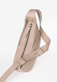 Borsa a tracolla in pelle beige con una tasca diagonale con zip nera e ampia tracolla regolabile su sfondo bianco.