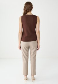 Kobieta stoi tyłem, ubrana w bezrękawnik w kolorze brązowym, beżowe spodnie i cieliste szpilki na białym tle.