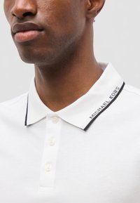 Gros plan d'une personne portant un polo blanc avec une patte de boutonnage à trois boutons et un bord de col étiqueté "MICHAEL KORS."