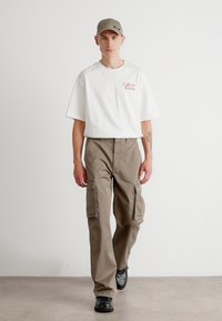 Hvid kortærmet t-shirt med rødt logo, brune cargo-bukser med sidelommer og sorte sko, fuldendt med en grå kasket.