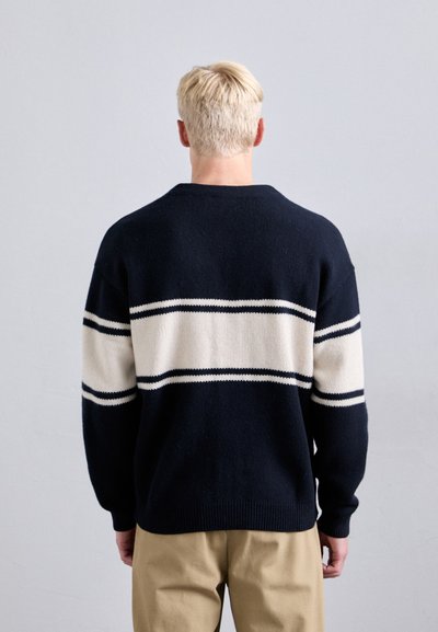 Ναυτικό μπλε πλέ knitted πουλόβερ με λευκές και ναυτικές ρίγες, στρογγυλή λαιμόκοψη, άνετη εφαρμογή και ριμπ μανσέτες, φορεμένο πάνω από μπεζ παντελόνια.