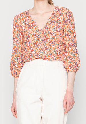 Femme portant un chemisier orange boutonné avec un motif floral coloré et des manches trois-quarts, associé à un pantalon blanc taille haute.