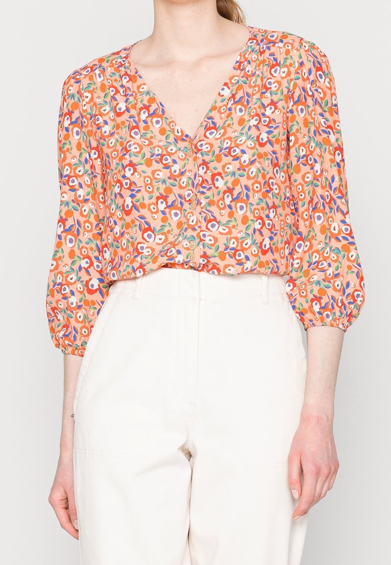 Femme portant un chemisier orange boutonné avec un motif floral coloré et des manches trois-quarts, associé à un pantalon blanc taille haute.