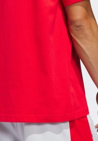 Højre arm på en person iført en rød skjorte og hvide shorts med røde striber, vist mod en lys baggrund.