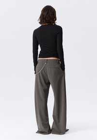 Une personne avec des cheveux mi-longs porte un haut noir à manches longues et un pantalon large à rayures grises avec un accessoire en chaîne, se tenant avec les mains dans les poches.