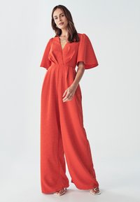 Mujer vistiendo un mono de pierna ancha de color naranja brillante con mangas cortas de vuelo y escote en V profundo, de pie frente a un fondo blanco liso.