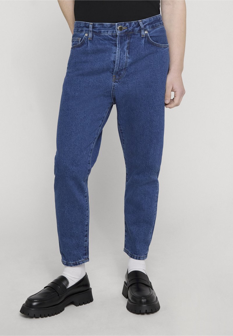Personne portant un jean bleu taille haute, des chaussettes blanches et des mocassins noirs épais, debout sur un fond clair uni.