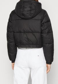 Veste matelassée noire courte avec capuche, présentant une texture matelassée, un ourlet élastique et un léger éclat. Portée par-dessus un pantalon blanc.