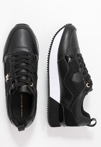 Baskets de sport noires en cuir lisse et perforé, avec une semelle en caoutchouc blanche, un laçage à l'avant et des accents métalliques.