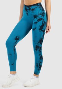 Kobieta w niebiesko-czarnych leginsach w stylu tie-dye i białych sneakersach, stojąca z lekko ugiętą jedną nogą na jasnoszarym tle.