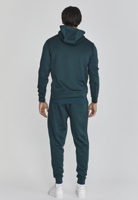 Sudadera color teal y joggers a juego, fabricados en un tejido suave y elástico con puños acanalados. Presenta un corte relajado y capucha ajustable.