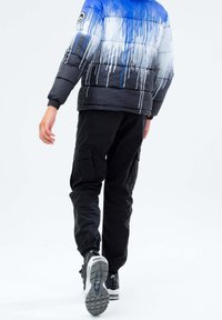 Persona caminando alejándose, vistiendo pantalones cargo negros, zapatillas negras y blancas, y un abrigo acolchado en gradiente azul, blanco y negro con un diseño de goteo de pintura.