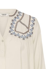 Blouse crème avec broderie géométrique bleue et marron sur l’empiècement, avec patte de boutonnage partielle et tissu froncé sous la poitrine.