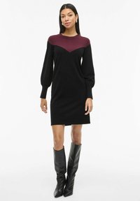 Czarna dzianinowa sukienka z bordowym karczkiem, długimi bufiastymi rękawami i prostym krojem. Noszona z czarnymi, skórzanymi kozakami za kolano.