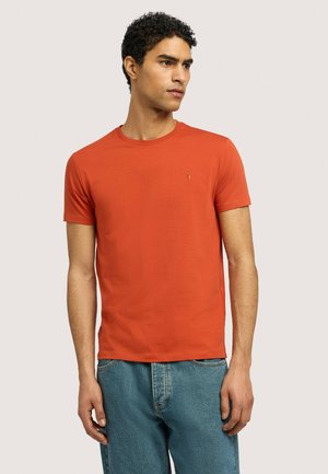 Mężczyzna w gładkiej pomarańczowej krótkiej koszulce i niebieskich dżinsach, patrzący w lewo na jasnym tle.