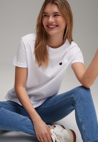 Witte katoenen t-shirt met opgerolde mouwen en zwart hartlogo op de borst, gecombineerd met blauwe spijkerbroek en witte sneakers met bruine accenten.