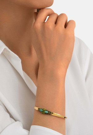 Bras portant un bracelet en or avec des pierres vertes et turquoise, reposant près du menton, habillé d'une chemise blanche.