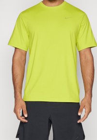 Hombre vestido con una camiseta deportiva de manga corta amarilla brillante de Nike y pantalones cortos atléticos negros, de pie con los brazos relajados a los lados.