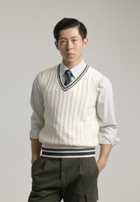 Chaleco de punto crema con cuello en V, con ribetes a rayas en verde oscuro y azul marino, combinado con una camisa a rayas en gris claro y blanco y pantalones en verde olivo.