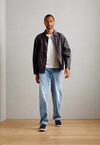 Black, button-up jacket over a white T-shirt, paired with light blue jeans and dark sneakers. Model stands on a wooden floor against a neutral wall.