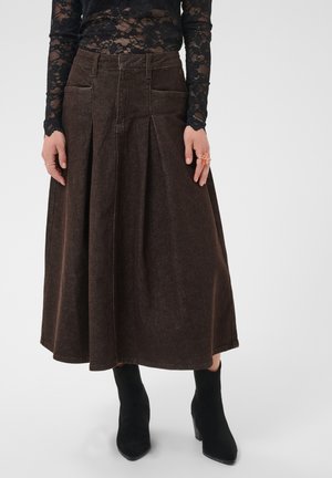 Femme portant un haut en dentelle noire transparente, une jupe midi marron évasée à taille haute avec des plis et des bottines noires, les mains le long du corps.