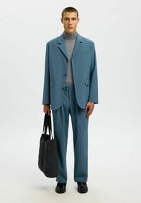 Svetlo modro lepo obleko z strukturiranim blejzerjem in ujemajočimi hlačami, nošeno čez sivo turtleneck. Model drži črno torbico.