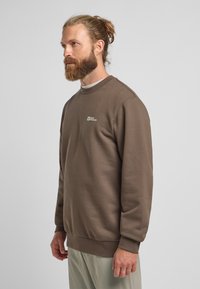 Felpa marrone realizzata in morbido tessuto, con scollo rotondo, maniche lunghe e un piccolo logo sul petto. Vestibilità leggermente rilassata.