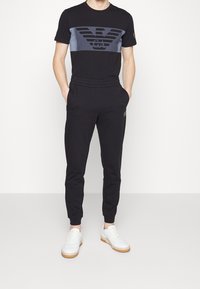 Svart t-shirt med en blå grafisk panel som har en logotyp, ihop med svarta joggers med elastiskt midjeband och vita sneakers med gummisulor.