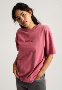 Camiseta de algodón rosa con mangas cortas, escote redondo y logo bordado en el pecho. Textura suave y corte holgado.