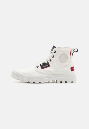 White canvas ankle boot with a textured rubber sole, featuring black and red accents, round logo patch, and lace-up design.