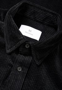 Sort corduroy skjorte med en struktureret overflade, der har en spread krave, sorte knapper og et hvidt mærke med "Matinique" logo.