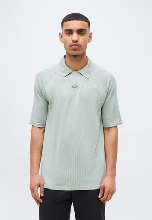 Νεαρός άνδρας που κοιτάει εμπρός, φοράει ανοιχτό πράσινο πουκάμισο πόλο με κοντά μανίκια και μαύρο παντελόνι, μπροστά σε απλό λευκό φόντο.