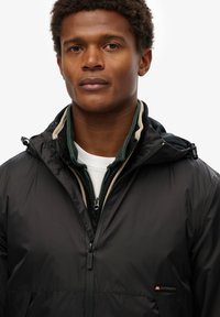 Zwarte waterdichte jas met een gladde textuur, verstelbare kap en opvallend logo. Gelaagd met een groene fleece en een witte shirt eronder.