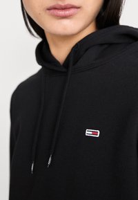 Svart hoodie med en framåtriktad känguruficka, justerbara snören och en liten broderad logotyp i vitt, rött och blått på bröstet.