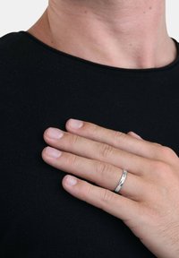 Eine Hand mit einem silbernen Ring liegt an einem schwarzen Shirt in der Nähe des Halses der Person und deutet auf eine Geste von Nachdenklichkeit oder Introspektion hin.