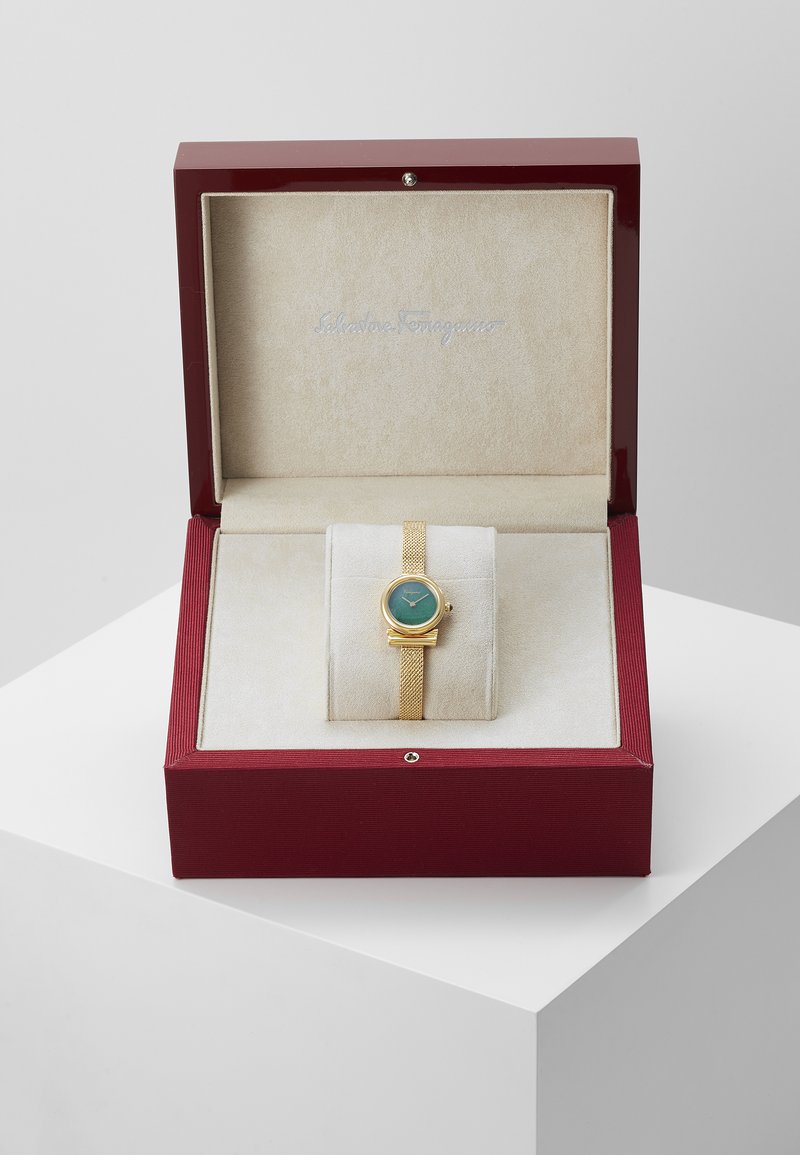 Montre dorée avec un cadran vert et un bracelet en maille, présentée dans une boîte bordeaux avec un intérieur en suede beige et le logo "Salvatore Ferragamo".