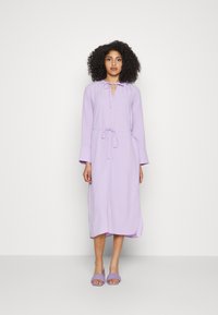 Vestido midi lavanda com mangas longas, cintura ajustável e decote aberto. Combinado com sandálias de slide abertas na frente em lavanda. Textura de tecido suave.