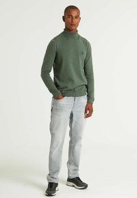 Grüner Rollkragenpullover aus Strickstoff, mit geripptem Saum und Bündchen. Kombiniert mit hellblauen Jeans und dunklen Sneakers.