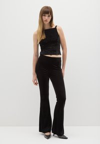 Giovane donna con capelli lunghi che indossa un crop top di velluto nero, pantaloni a vita alta svasati coordinati e tacchi alti neri con borchie.