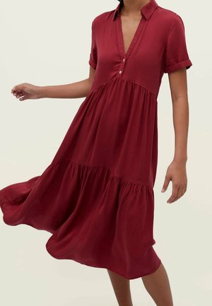 Femme portant une robe midi fluide rouge foncé à manches courtes, avec un col et un décolleté en V boutonné, en plein mouvement sur un fond uni.