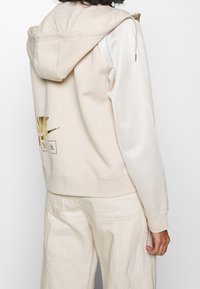 Beige hoodie med huva, långa ärmar, kontrasterande vita sidopaneler och en gyllene grafik på ryggen som har texten "Just Do It".